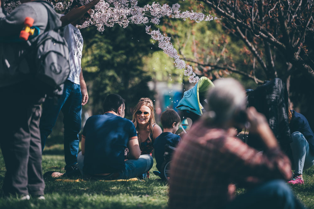 A young woman, man and child sit on the gound in a prk with others around them