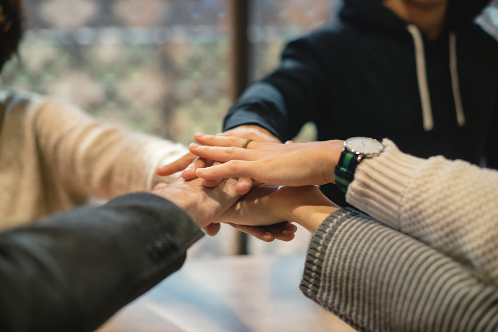 Five individuals in business casual attire place their hands in a "Go Team" formation