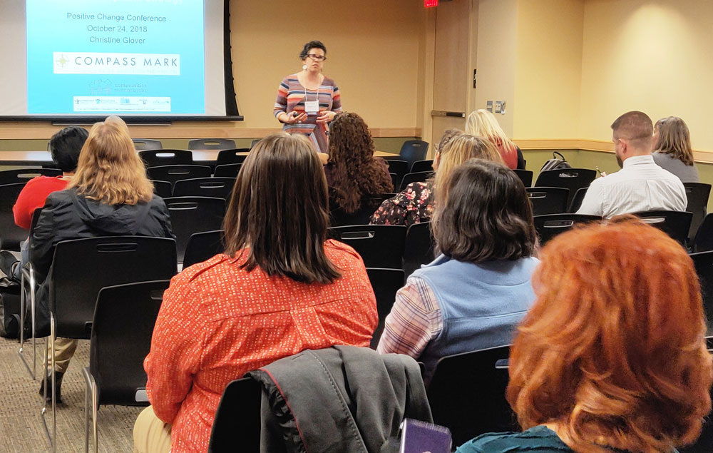 A woman presents to a group of people at a conference in front of a projection screen.