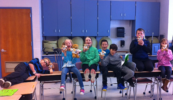 Seven elementary-aged students sit on desks in their classroom, holding stuffed bears and grinning.