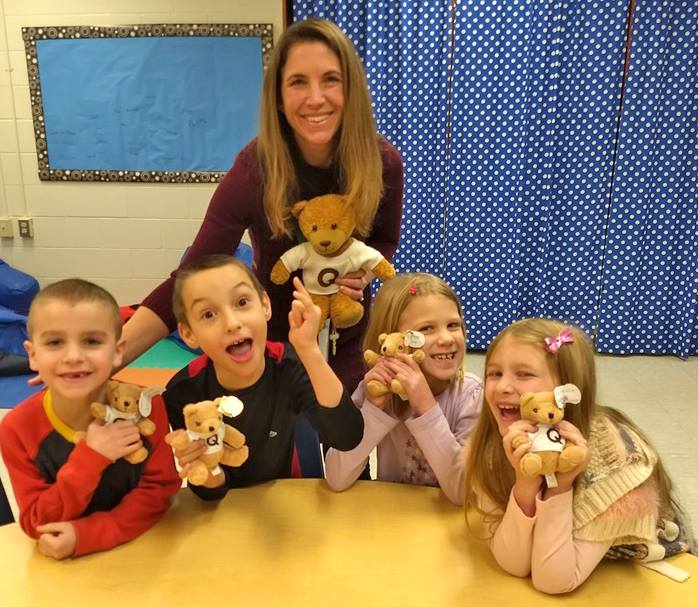 A young teacher and 4 smiling elementary-aged kids smile and hold up stuffed teddy bears