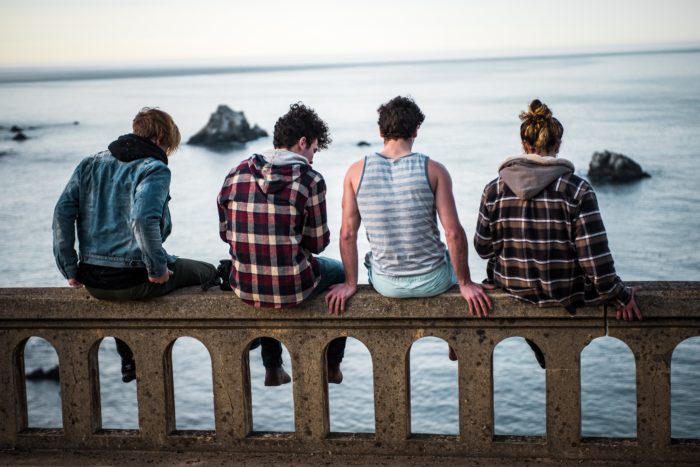 We see the backs of 4 teens sitting on the edge of a bridge- water in the background. It's gray out, and they seem quiet.