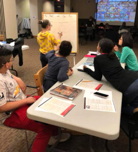 Five people work out a problem on a white board- we see their backs around a table.