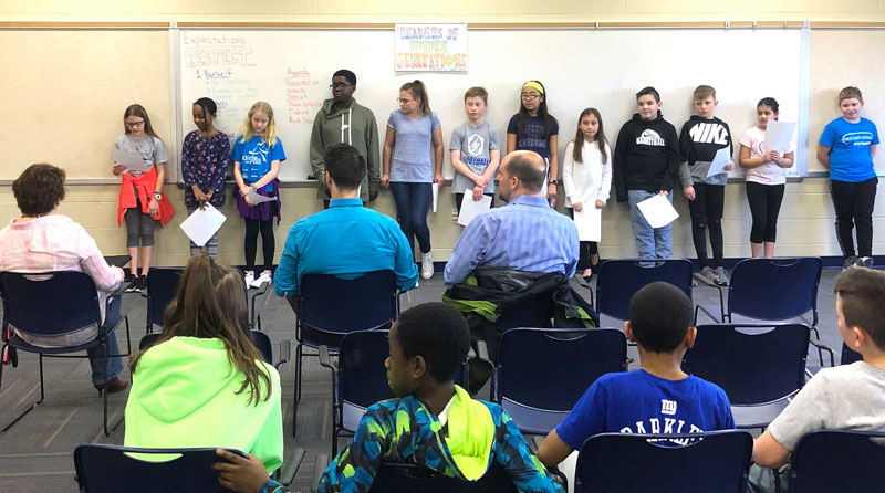 A group of middle school students present an idea to a group of adults. The kids stand at the front of the room, and we see the backs of the adults, seated.