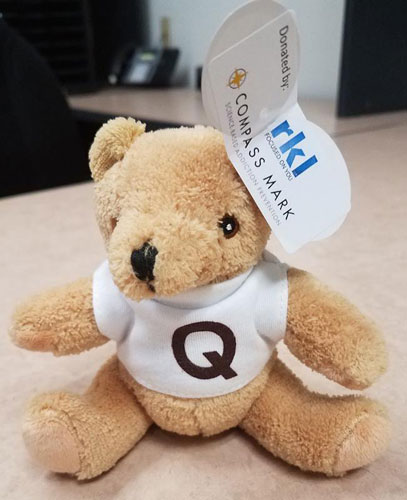 A teddy bear wears a white shirt with a "Q" on it, as well as a tag that reads, "Sponsored by RKL"