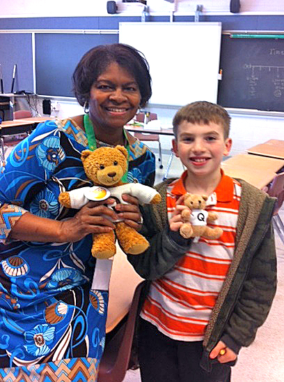 In a classroom, a woman and elementary-aged student pose with a stuffed bear.