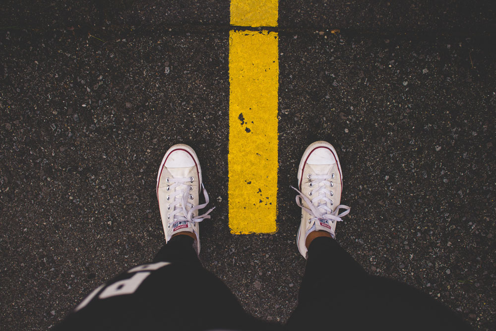 We see 2 feet in white sneakers, one on either side of a yellow dotted line on a roadway.