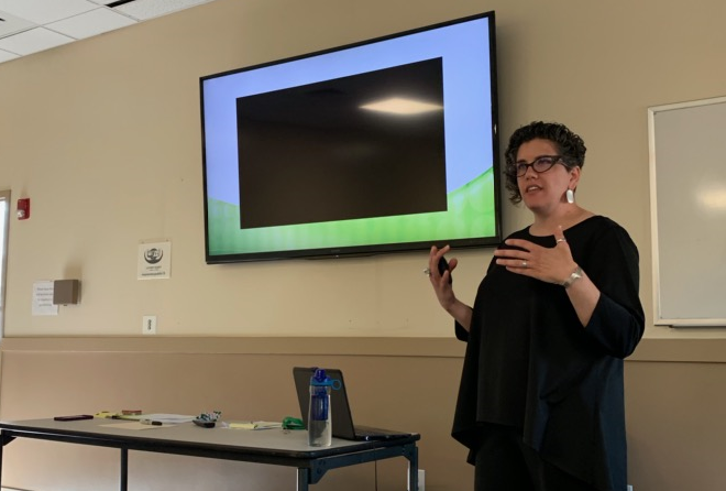 A woman with dark hair and shirt stands in front of a projection monitor, obviously speaking to an audience.