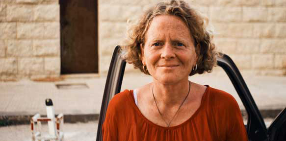 A middle-aged woman in a rust shirt and gold necklace is seen close up- maybe smiling, maybe squinting.