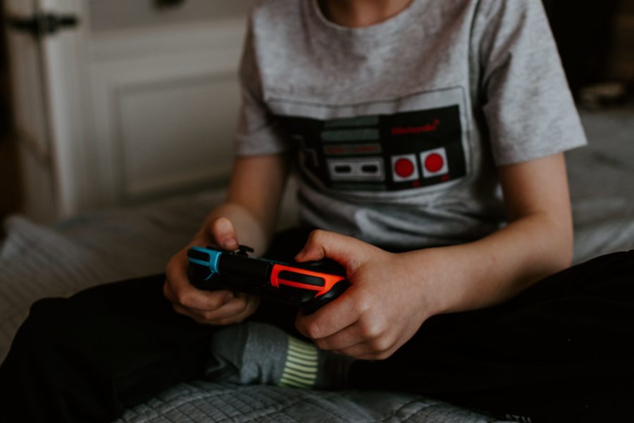 We see the torso and legs of a young person, possibly male or gender-neutral, sitting on a bed and playing video games.