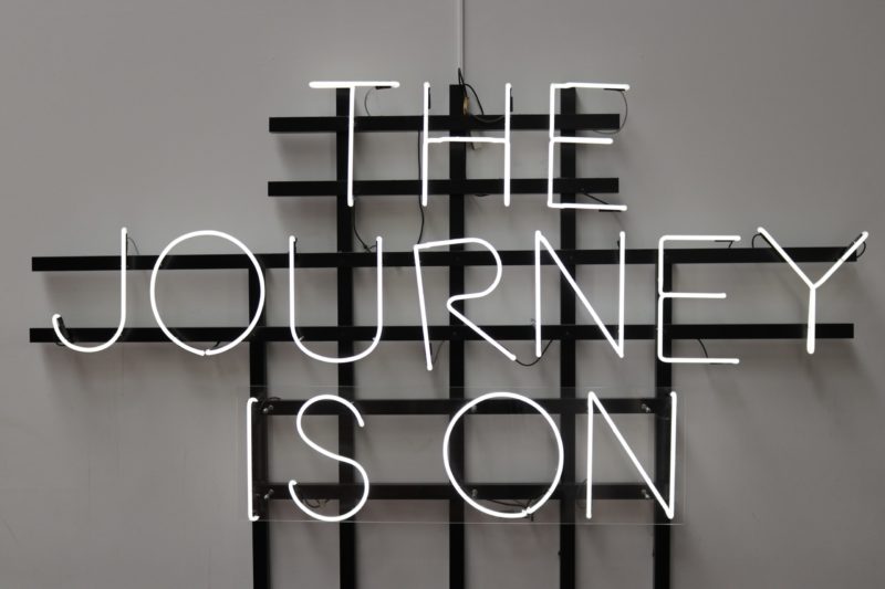 A neon sign is fixed to a white wall. The letters are white, and read: The Journey is On.