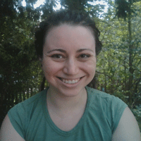 Close up of a young woman smiling in a casual shirt with green trees behind her.