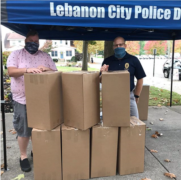 Two men stand outside under a tent in back of cardboard boxes. They are wearing masks.