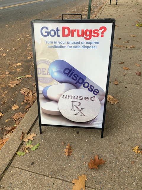 An outside sign on a sidewalk says, "Got Drugs?" with tablets and pills. 