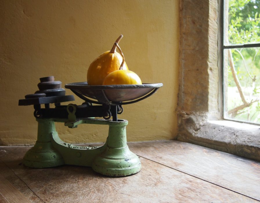 On an old wooden table we see an old-fashioned balance scale, with pears and weights in the pans. It's near a window.