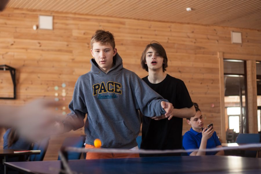 Two teen boys are shown playing ping pong in a rustic hall.