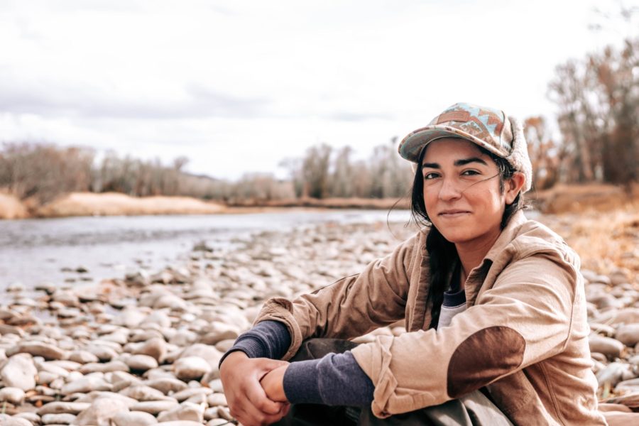 A woman in her 20's or 30's is sitting on the rocky shoreline of a river. The rocks are round and smooth. She is dressed in coat and cap to stay warm.