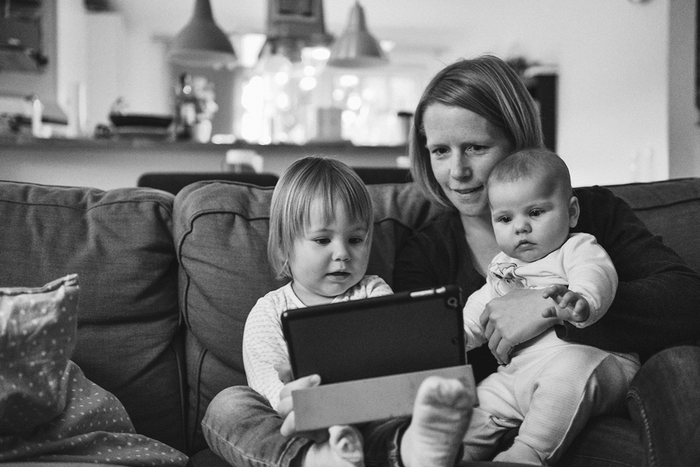 Black and white image shows a mom at home on a couch with a baby and a toddler in her lap. They're all looking at a tablet.