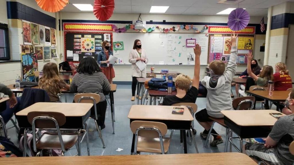 We look toward the front of a middle-school classroom at the adult leaders standing at the front, masks on. We see from behind students raising their hands.