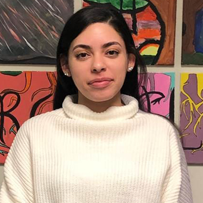 A young woman in business casual attire stands in front of colorful artwork.