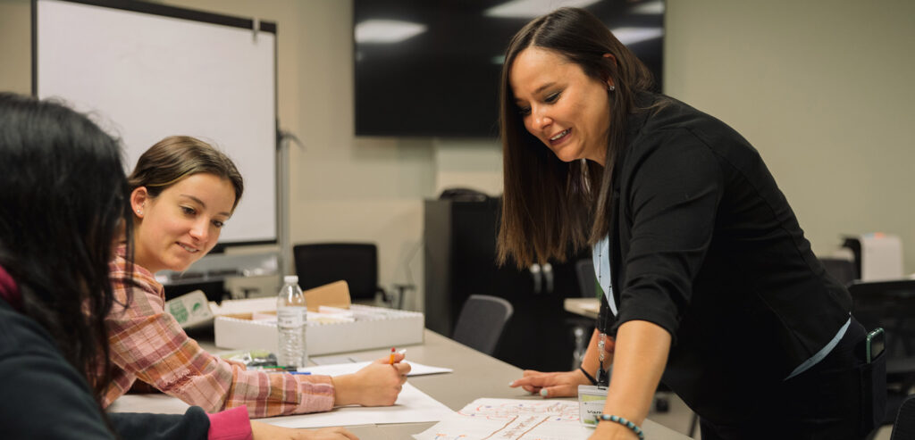 A woman leads a workshop with a white board behind her. She leans over some attendees' table to assist them.