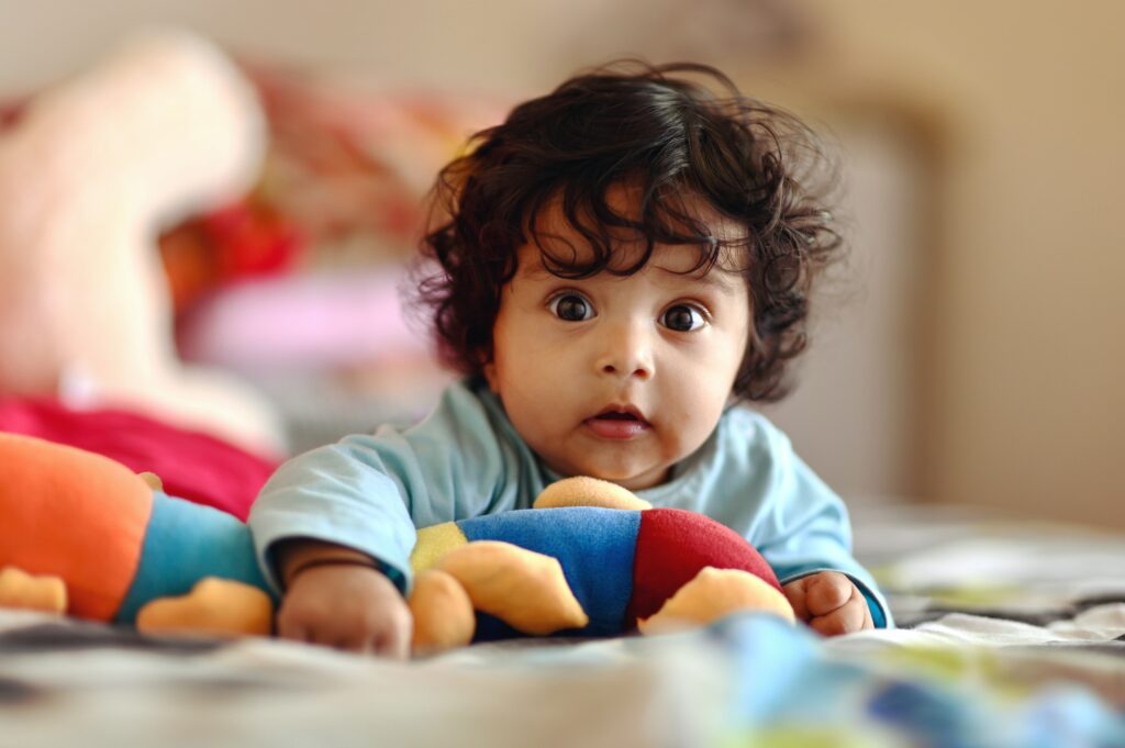 We are at eye level with a baby who is laying on his tummy on the floor. He's playing with colorful toys and has a head of curly, dark hair. 