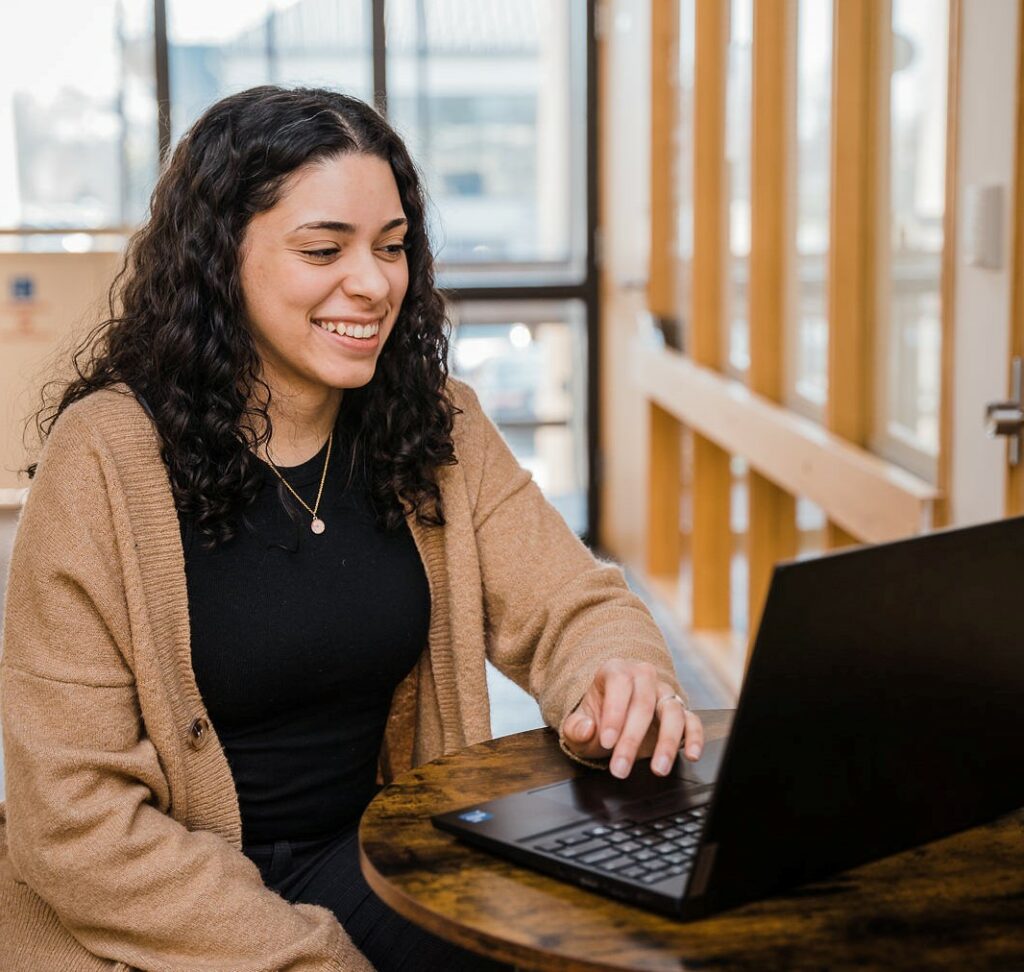 A woman in business casual clothing is sitting at a laptop, engaged and smiling.