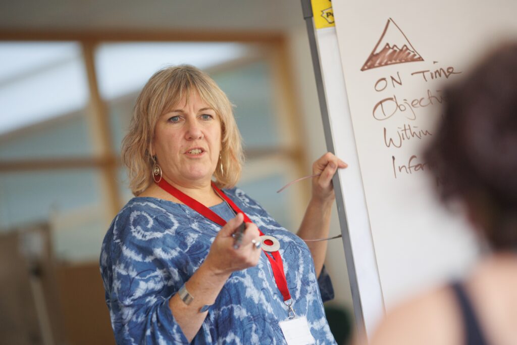 A woman stands at the front of a room next to a white board and appears to be speaking