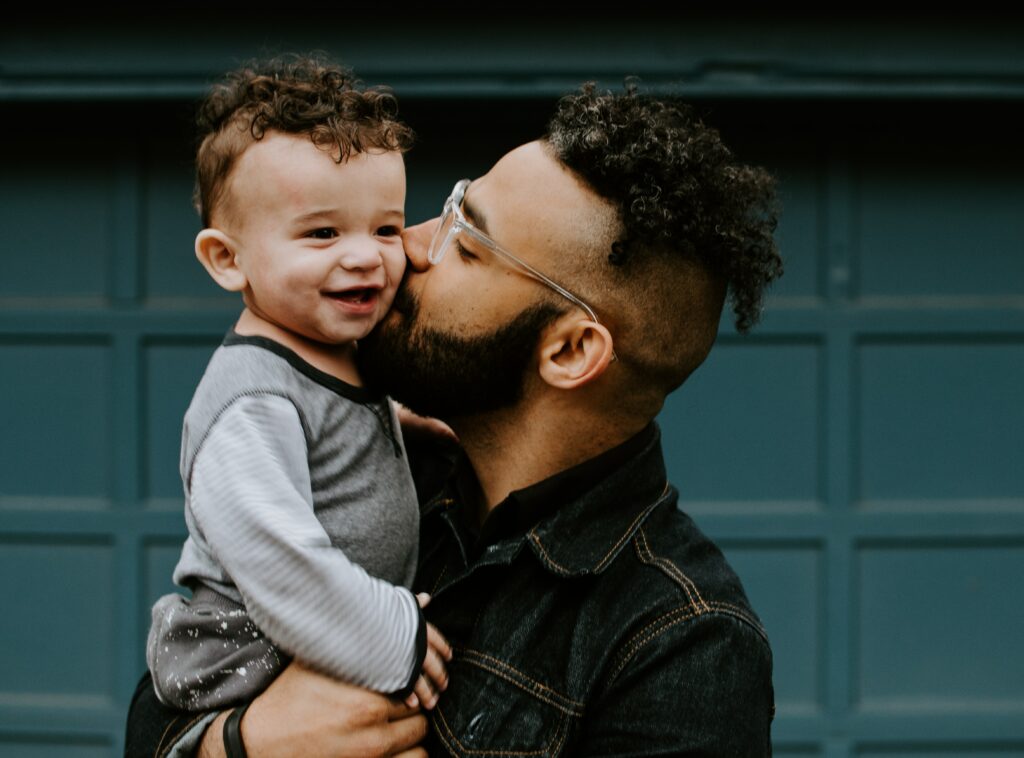 Closeup of a young man holding a toddler and giving him a kiss on the cheek.