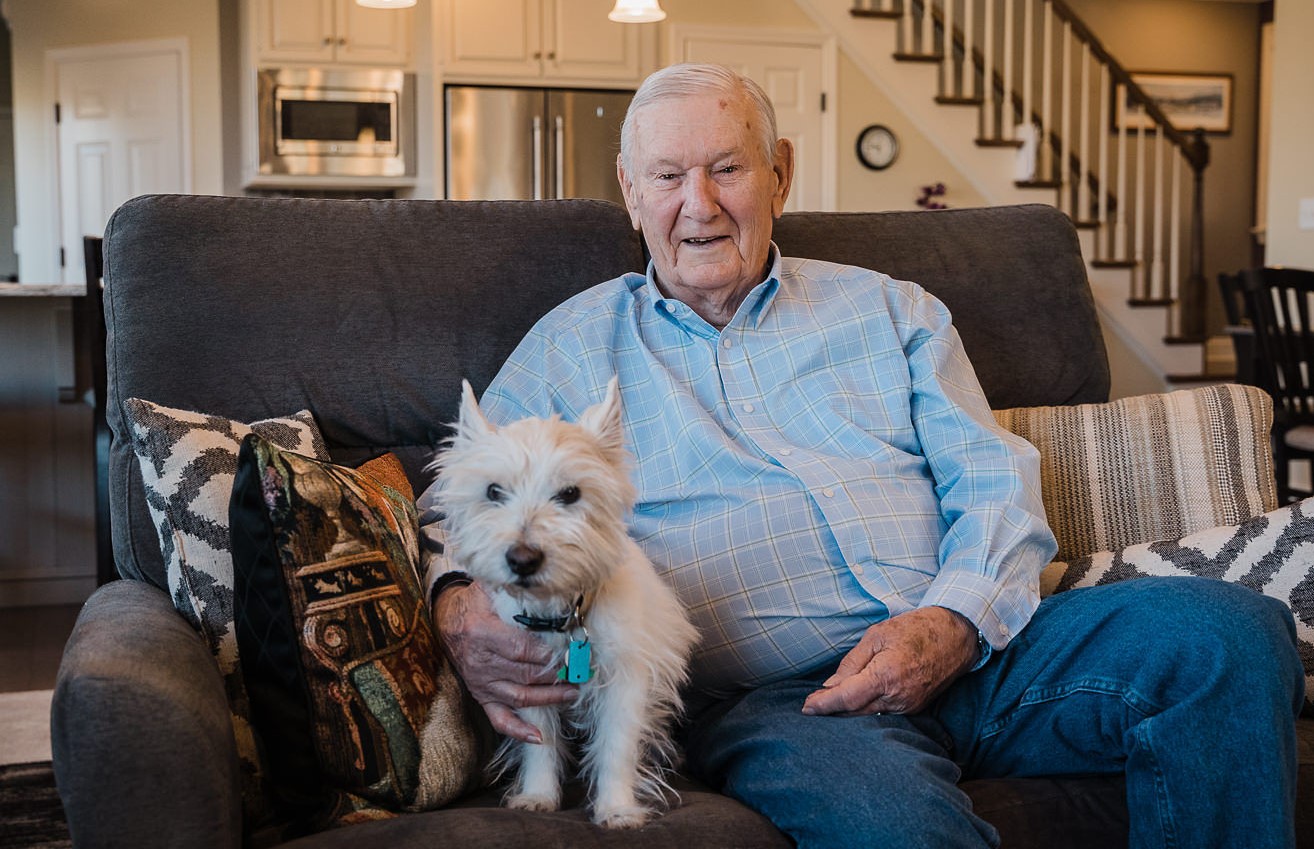 An older man is dressed in an oxford shirt & jeans, sitting on his couch in a nice, modern living space. His fluffy, white dog sits with him.