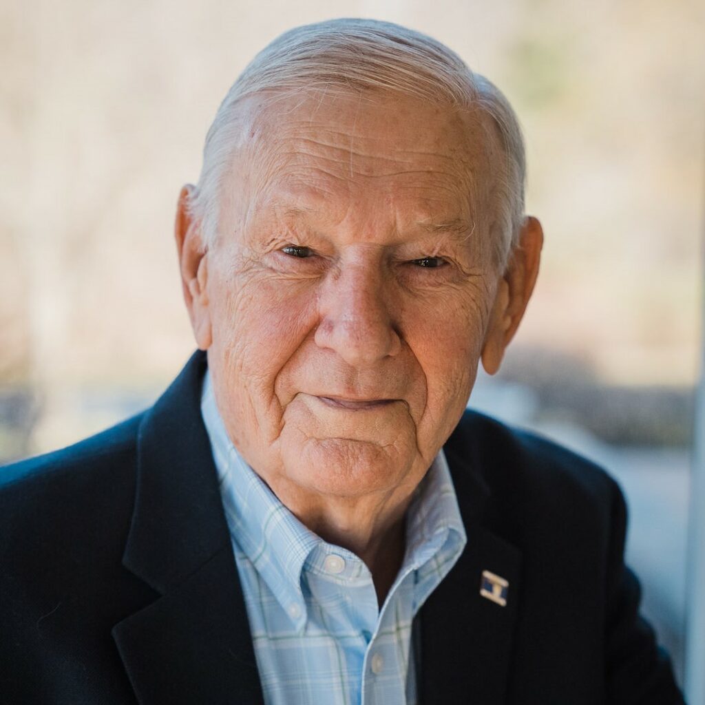 Closeup of an older man. He wears an oxford shirt and dark blazer and looks kindly at the camera. 