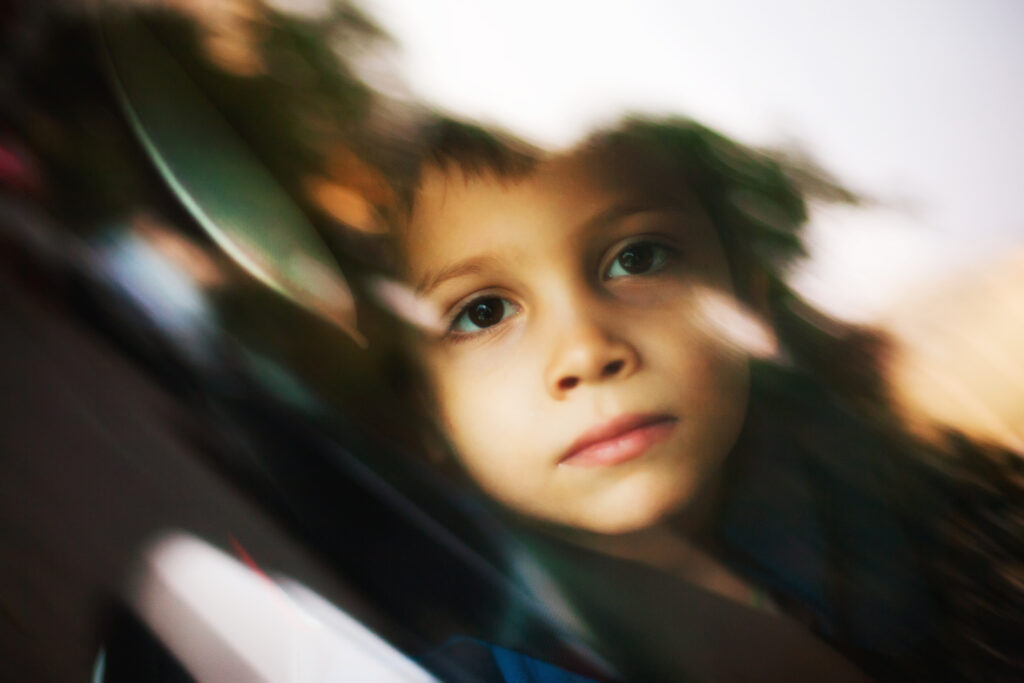 A child behind glass looks slightly blurred. We see the reflection of trees on his face. He looks serious. He's around 5 years old.