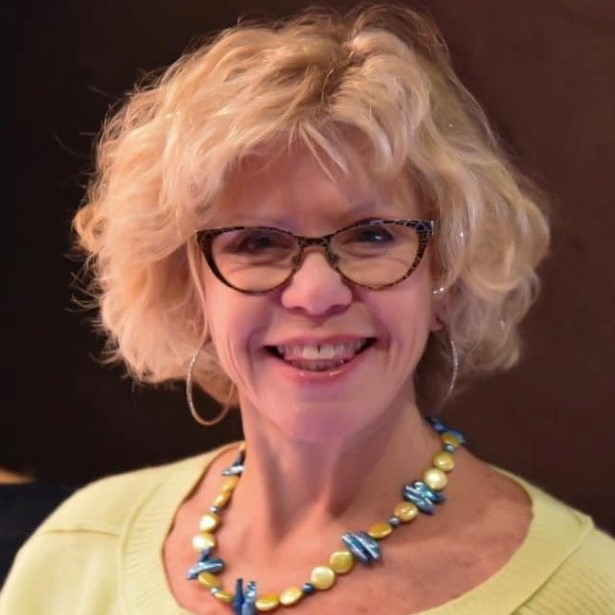 Marilyn G. Stein has short wavy hair with some sparkles in it, and is wearing a yellow shirt with a colorful necklace. She is smiling. 