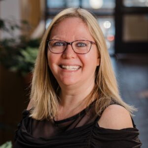 Headshot of a woman in black business casual attire in an atrium with lots of windows