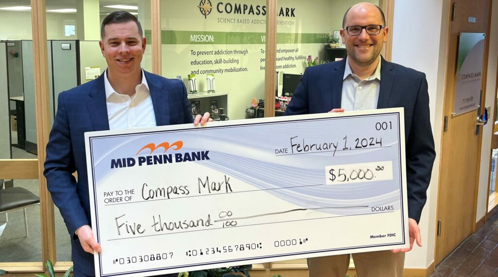 Two men hold a giant check made out for $5,000 in the atrium of a building