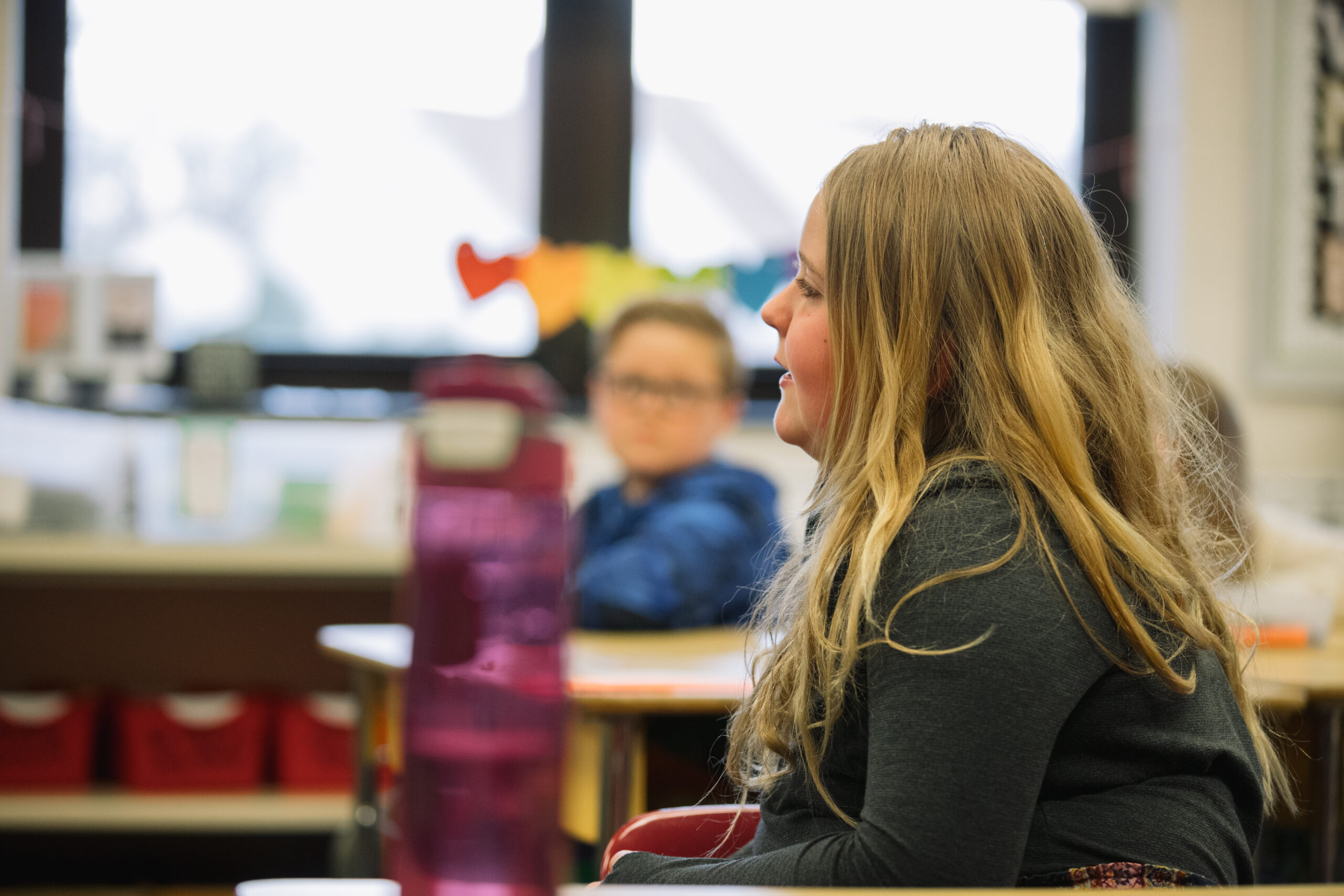 In a classroom, we see a little girl from the side. She is getting ready to answer a question. 