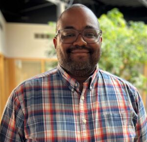 Headshot of a man in business casual clothing, in an indoor atrium with plants, smiling