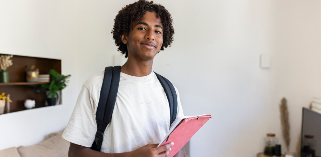 A teenage boy stands in a bedroom, getting ready for school. He has his backpack on, and is holding a notebook.
