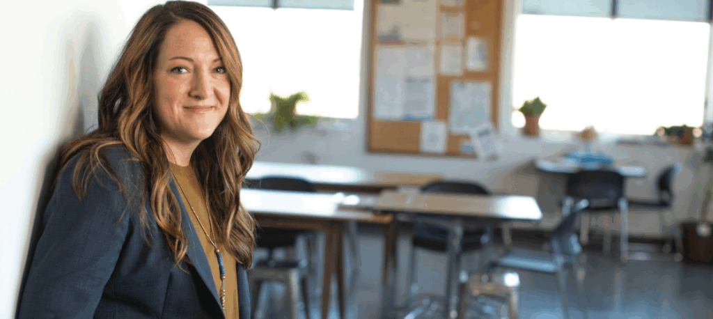A teacher leans against a wall in her classroom with desks in the background