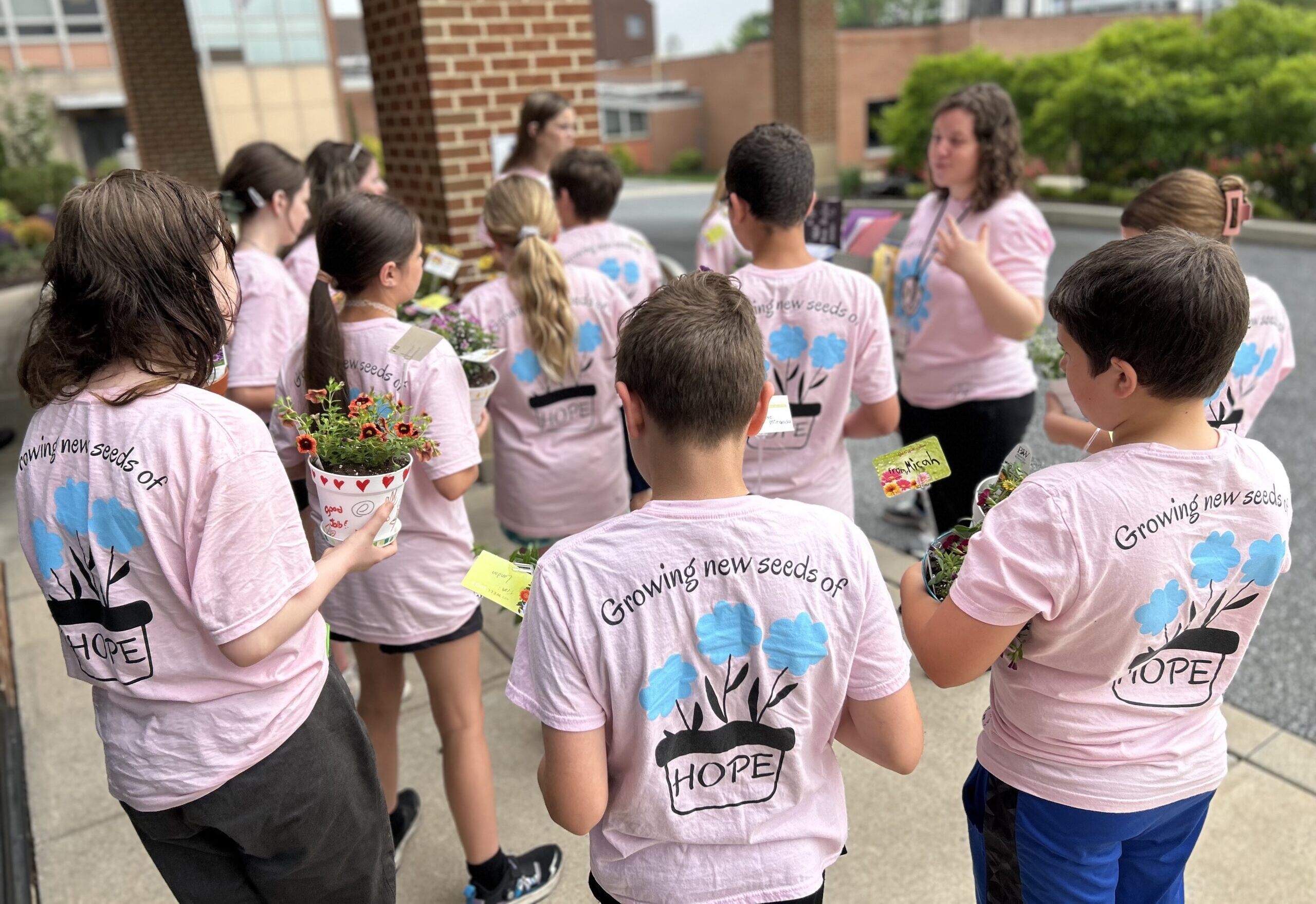 We see the backs of about a dozen middle-school students, who all have t-shirts that say, The Positive Planters. They are facing their adult teacher, who has the same shirt. They are outside.