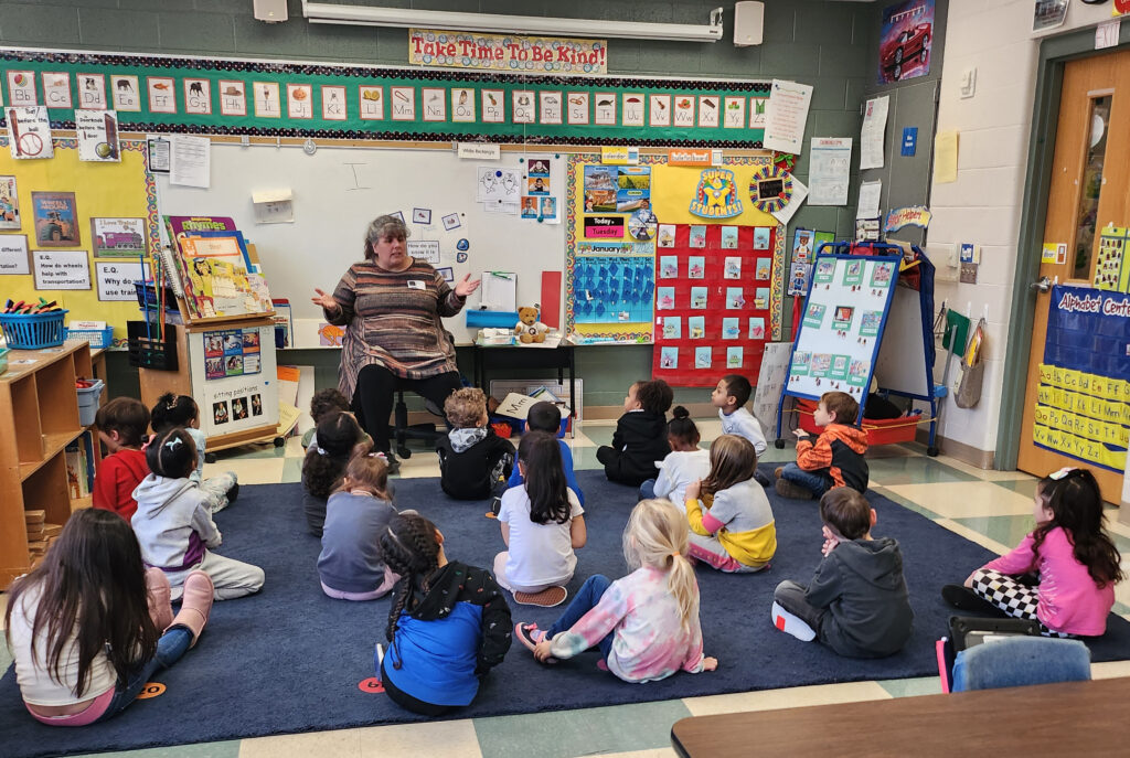 A teacher sits in front of a classroom of little children, who are sitting on the floor. 