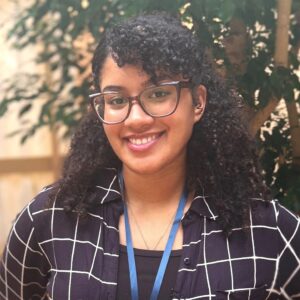 Headshot of a young woman in business casual clothing with a lanyard. She's in an atrium in with fig trees behind her.