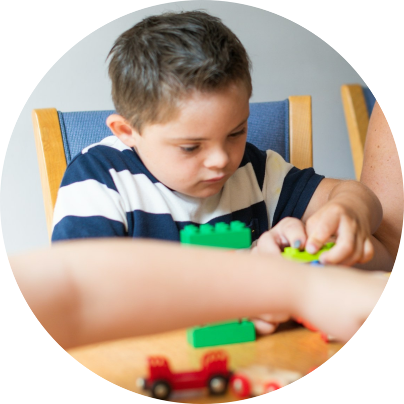 Closeup of a small boy seated, playing with toys at a table. At least one adult is present, reaching across in front of him.