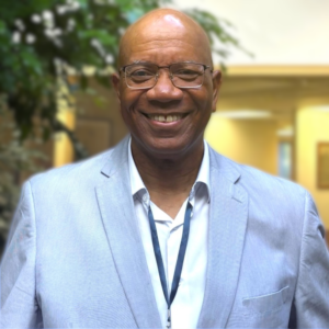 A man in collared shirt and light-blue jacket stands smiling in an indoor atrium with trees. He is surrounded by natural light and he is smiling.