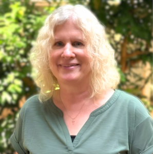 Headshot of a woman in casual clothing. She's standing inside the atrium of an office building in the morning, and light is scattered over the indoor fig trees behind her.