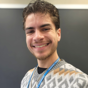 A young man in a sweater and lanyard smiles in front of a blackboard.