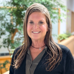 A woman in business casual attire stands flooded in natural light. She's inside an office building with skylights and large ficus trees grow behind her. She's smiling.