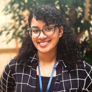 Headshot of a young woman in business casual clothing with a lanyard. She's in an atrium in with fig trees behind her.