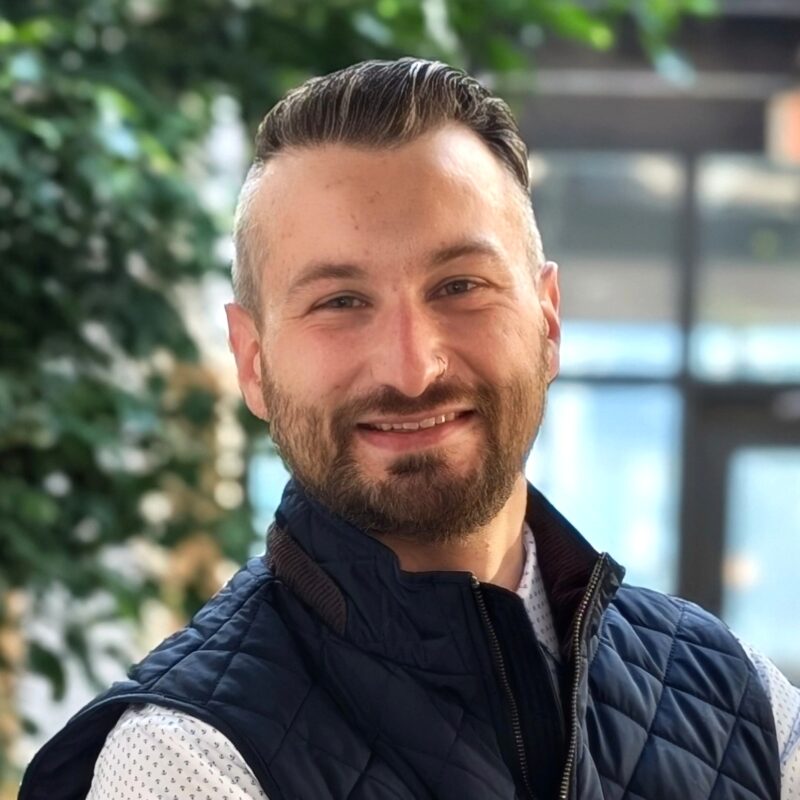Headshot of a man in an atrium inside a business park. We see indoor trees. He has on a quilted vest, collared shirt, and watch.