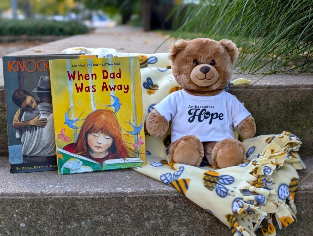 Outside on concrete steps, we see a cozy yellow blanket with bees on it, a teddy bear with a white t-shirt on that says Ambassadors for Hope, and two books- one called When Dad Was Away, and one called, Knock Knock.